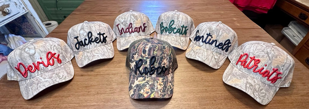 Lineup of puff embroidered camo hats for Robertson County schools — Devils, Jackets, Indians, Bobcats, Sentinels, Patriots, and RobCo trucker hat