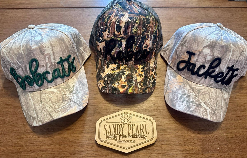 Three Sandy Pearl camo hats side by side — Bobcats, RobCo trucker, and Jackets — with Sandy Pearl branded wood plaque
